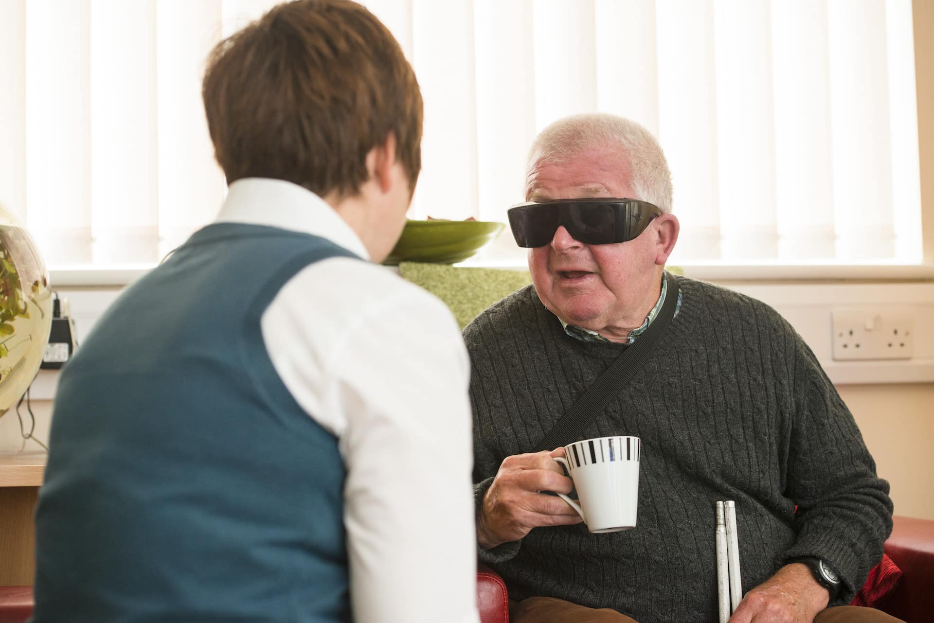 A man with dark glasses, a cup of tea and a folded up cane sits down talking to a lady in the foreground.