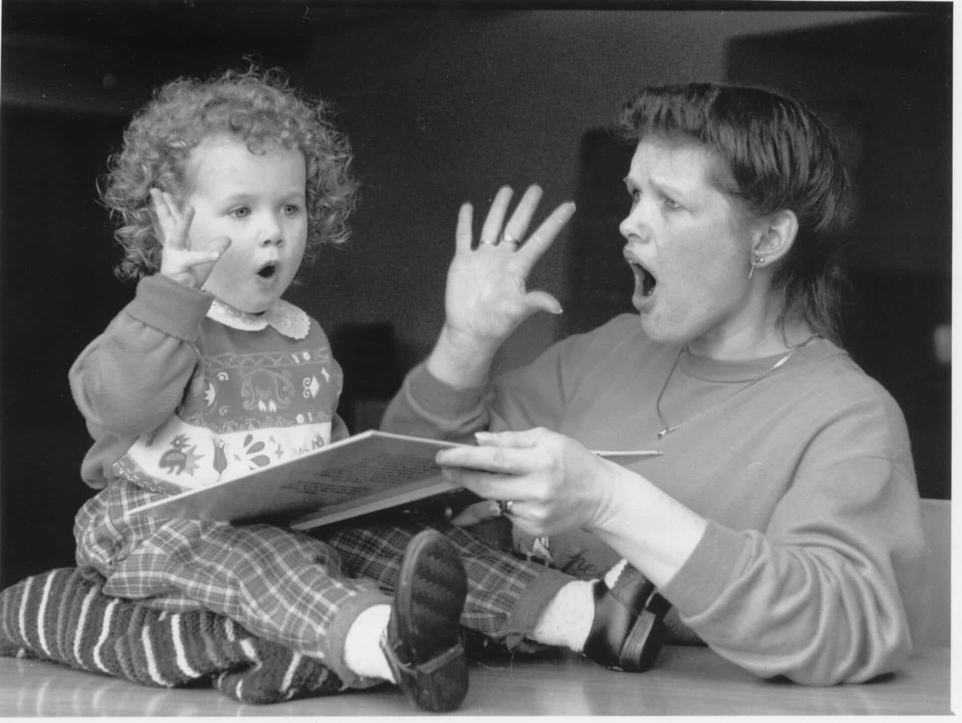 Mother with her young daughter reading a book and doing the sign for orange. Black and white image.