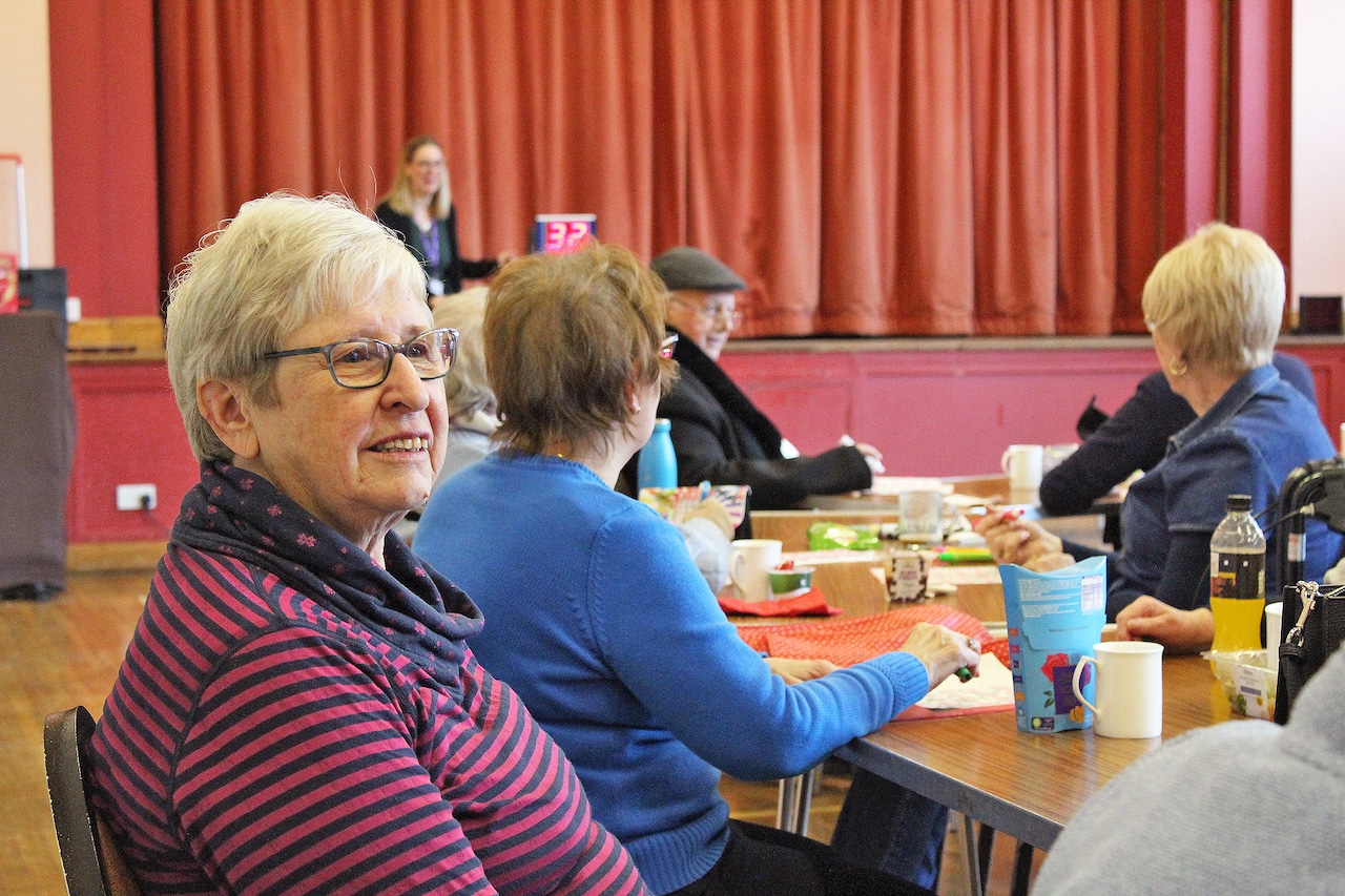 Several people sit around tables with drinks and snacks playing Bingo with a lady in the foreground smiling at someone off camera.