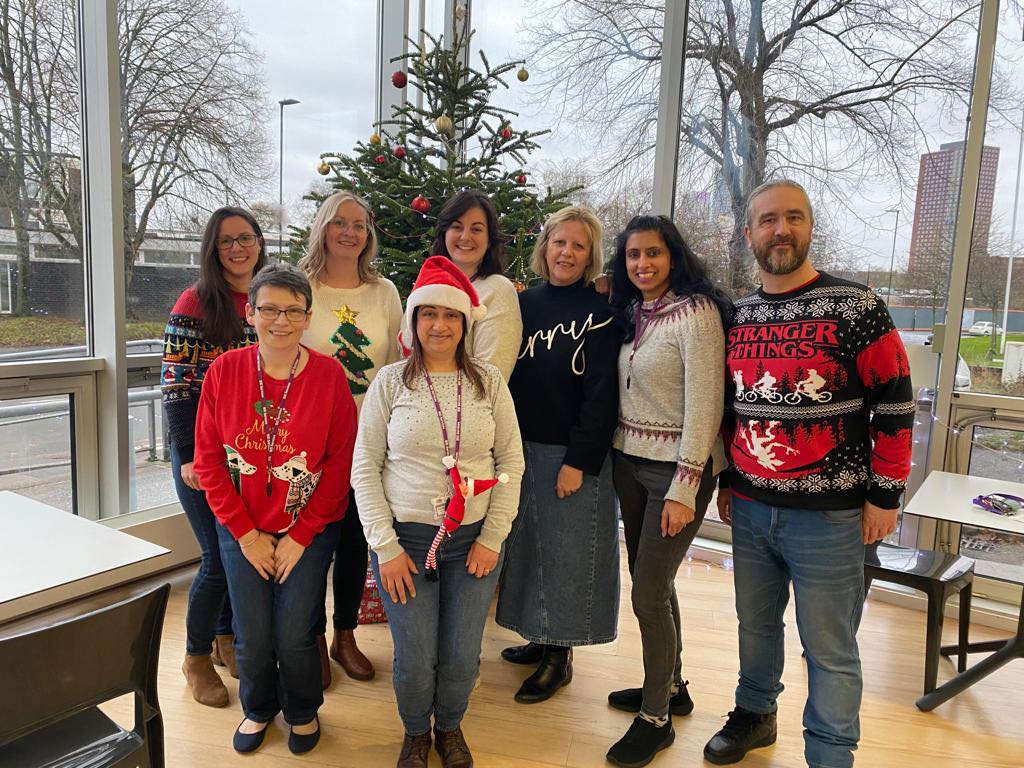 A group of 7 women and 1 man stood in front of a large Christmas tree. They're all wearing Christmas jumpers and smiling at the camera.