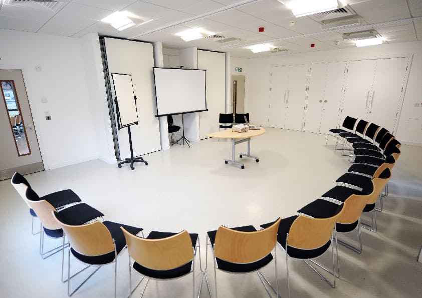 Bright room with white walls and flooring. The chairs are set up in a semi circle., all facing a blank presentation screen.