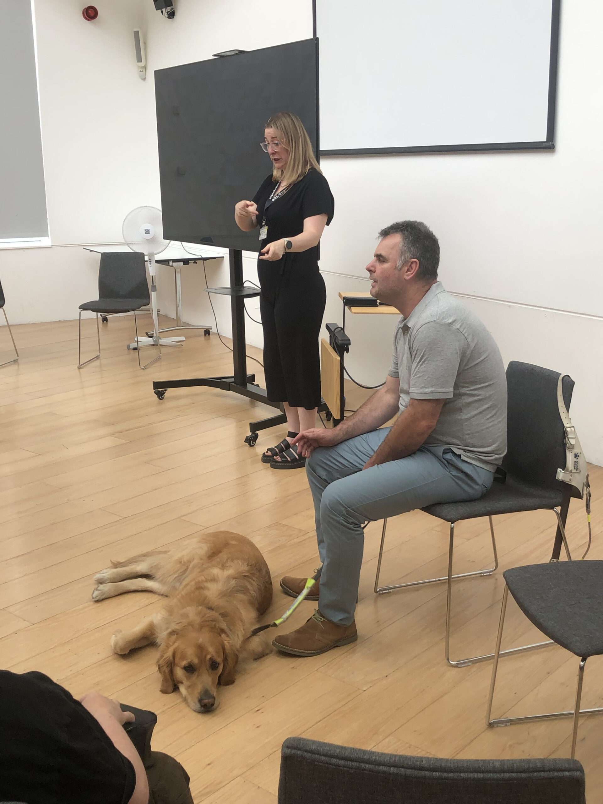 Alex is seated at the front of a room with his guide dog, Ned laying by his feet. He is talking to people off-camera. Beside him is a female BSL interpreter.