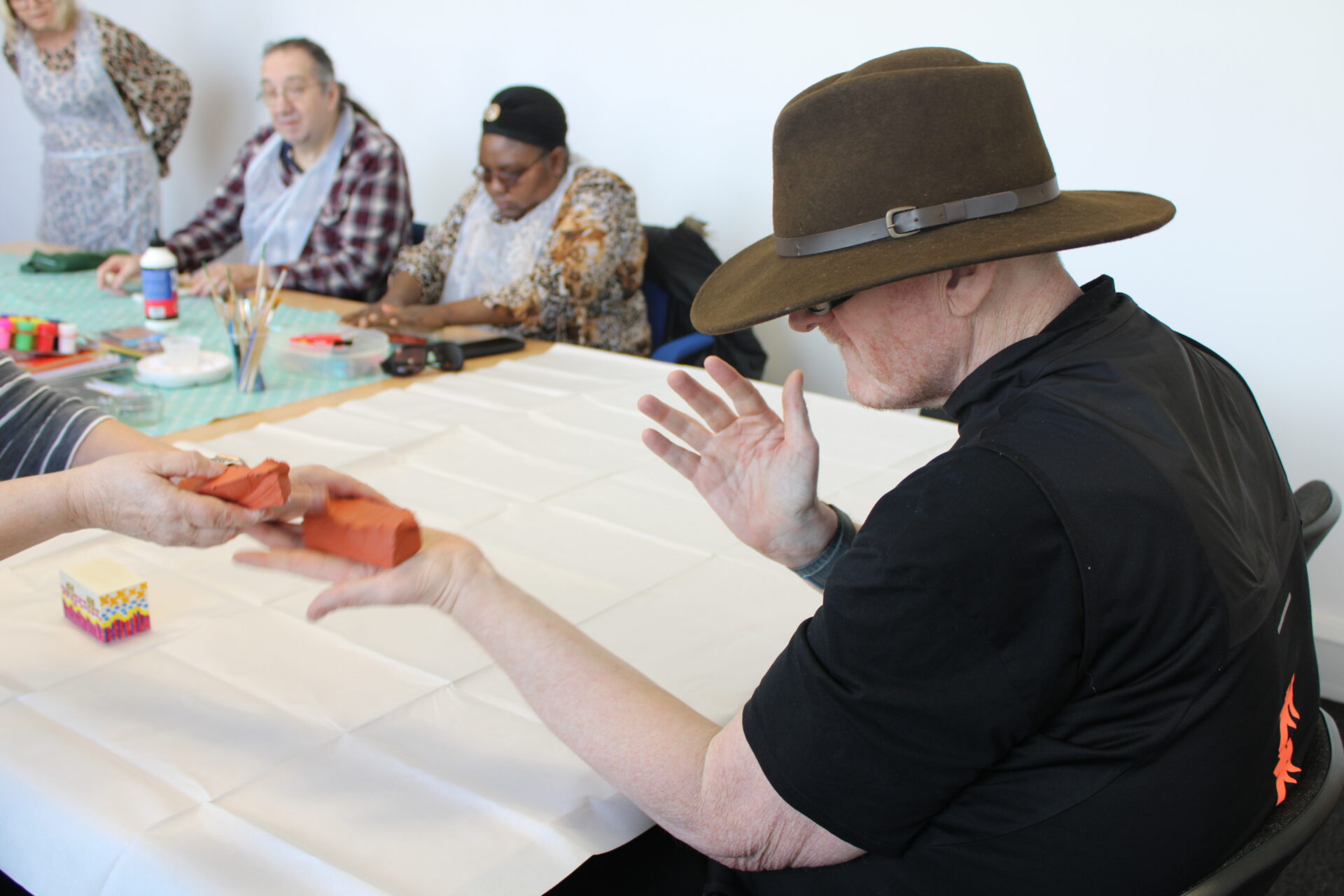 A man wearing a brown cowboy hat is sat at a table and being handed some orange clay. 