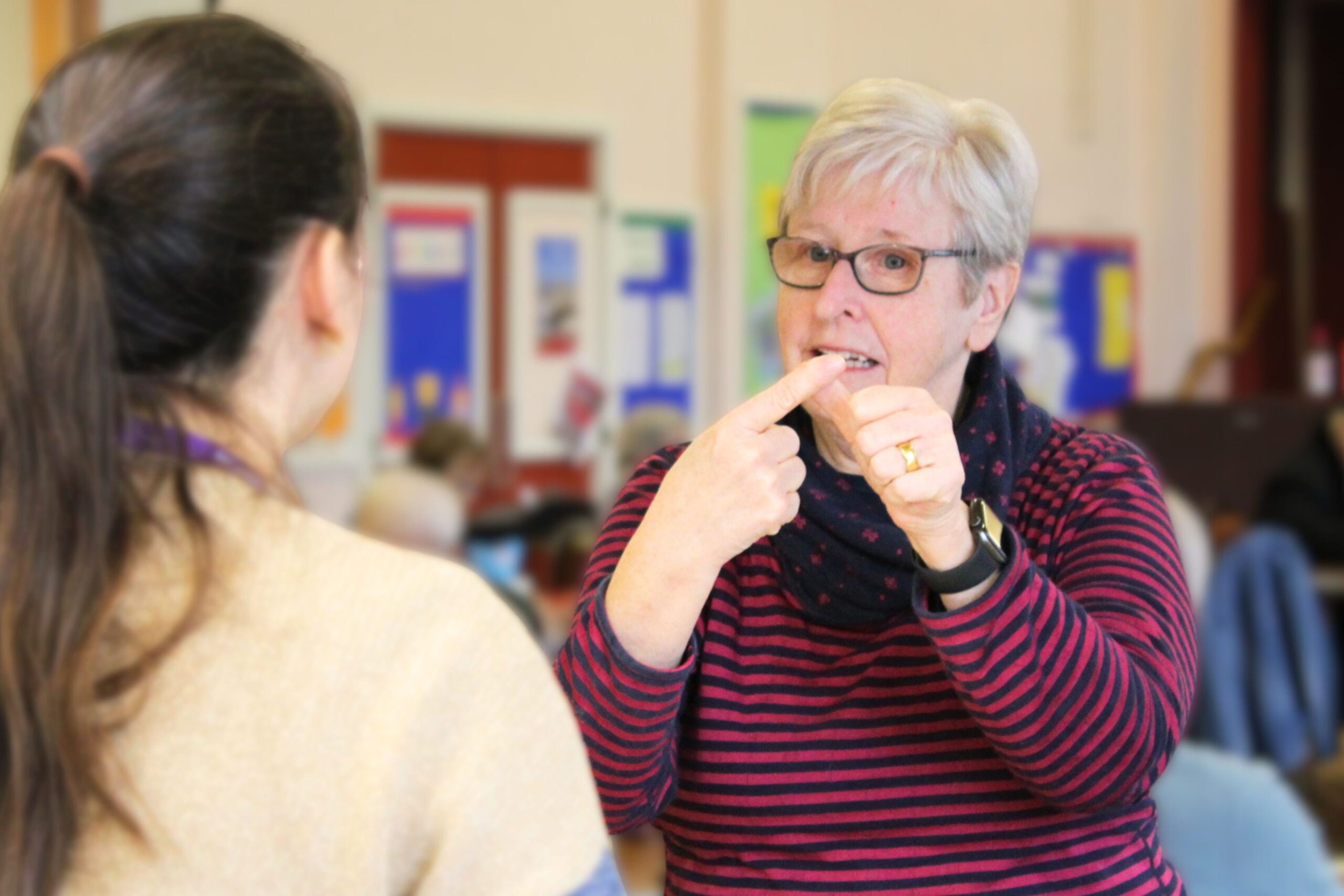 An elderly white woman is stood using British Sign Language to someone in the foreground.