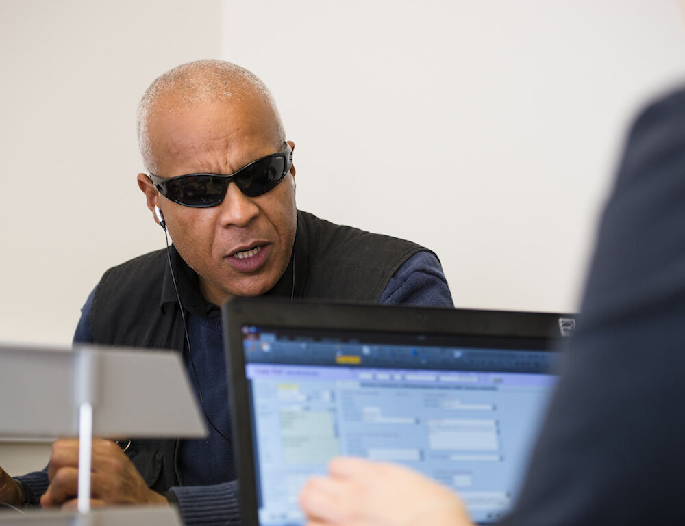 Asian man with dark glasses talking to a lady working on her laptop